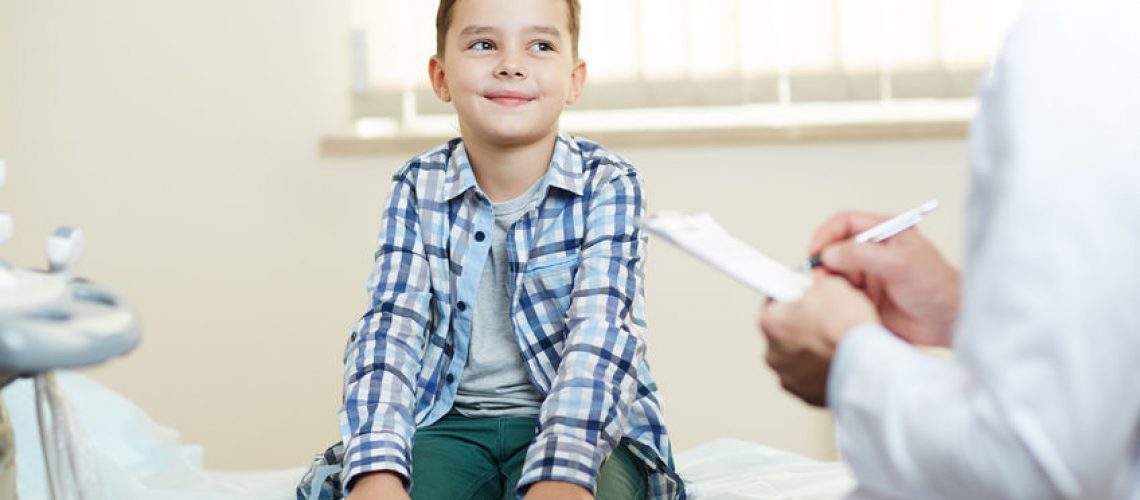 Happy little boy looking at his doctor while he making prescriptions and notes in medical form